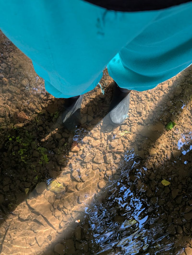 black wellingtons standing in a shallow stony brook, clear water running over the toes, teal waterproof trousers above